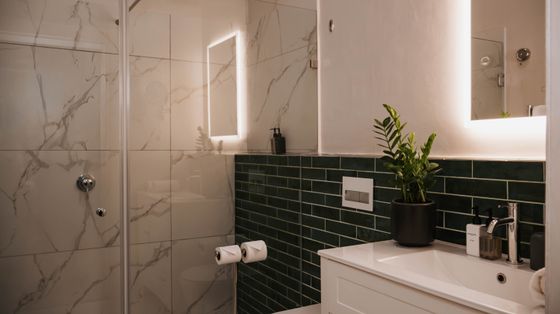 Bathroom with a glass-walled shower, marble-look tiles, dark green subway tile, a white vanity with a plant, and backlit mirrors.