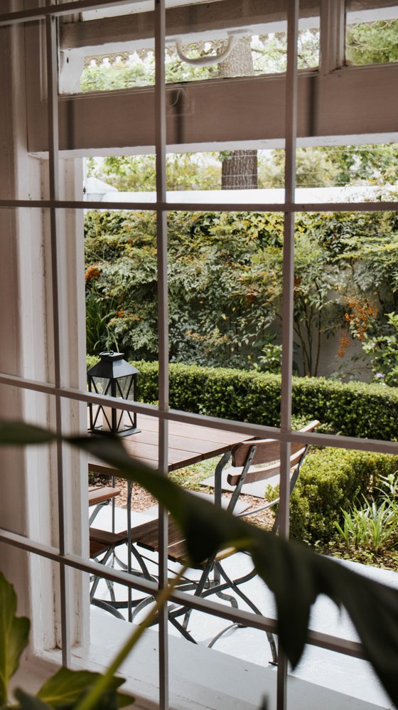Through a paned window, a wooden table with metal chairs sits in a garden courtyard beside a black lantern and a clipped hedge.
