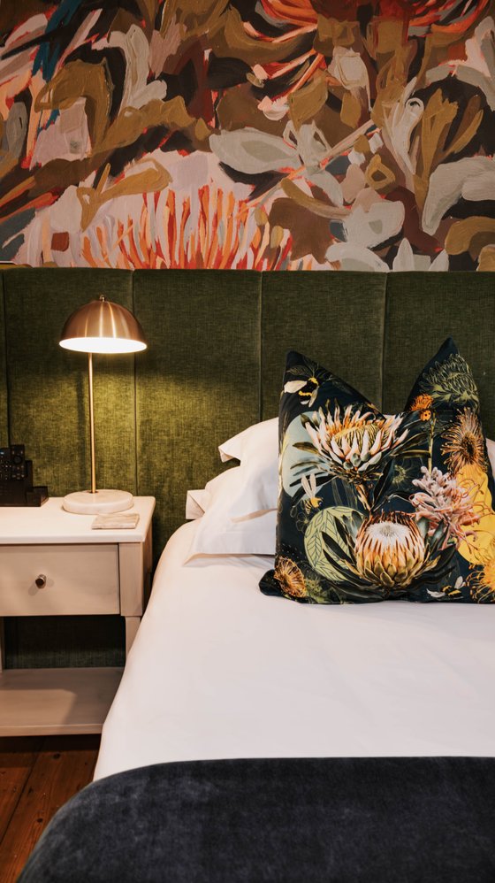 Bed with white linens, dark green upholstered headboard, floral throw pillows, and a brass lamp on a bedside table beneath a painted floral mural.