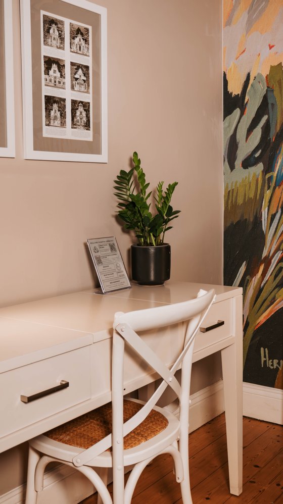 White desk with a cross-back chair and a potted plant beside framed black-and-white architectural prints and a colorful painting.
