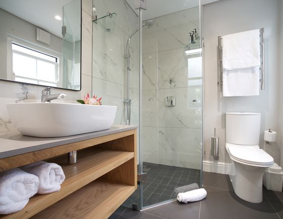 Bathroom with a white vessel sink on a light wood vanity, a glass-walled marble-tiled shower, a white toilet, and rolled white towels.