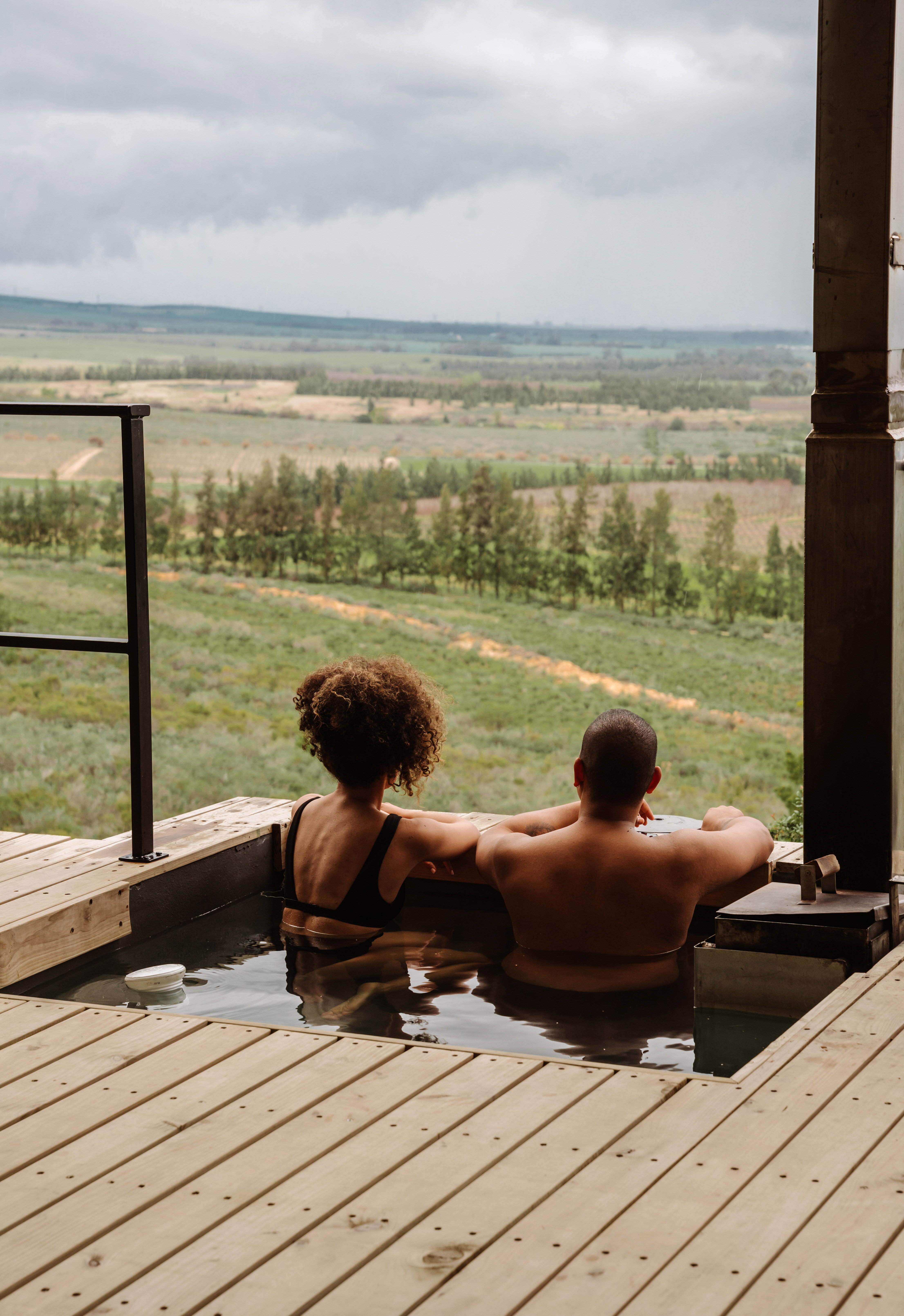 The Outpost at Bergsicht Country Cottages: A romantic couple's breakaway in the Tulbagh valley. The image shows a man and woman soaking in a private wood-fired tub—which can be heated for winter or kept cold as a plunge pool in summer—set against a backdrop of rolling vineyards and distant mountains under a dramatic sky.Tulbagh accommodation  Romantic getaway Western Cape  Wood-fired hot tub cottages  Self-catering Tulbagh  Couples retreat South Africa  The Outpost Bergsicht  Wine farm views