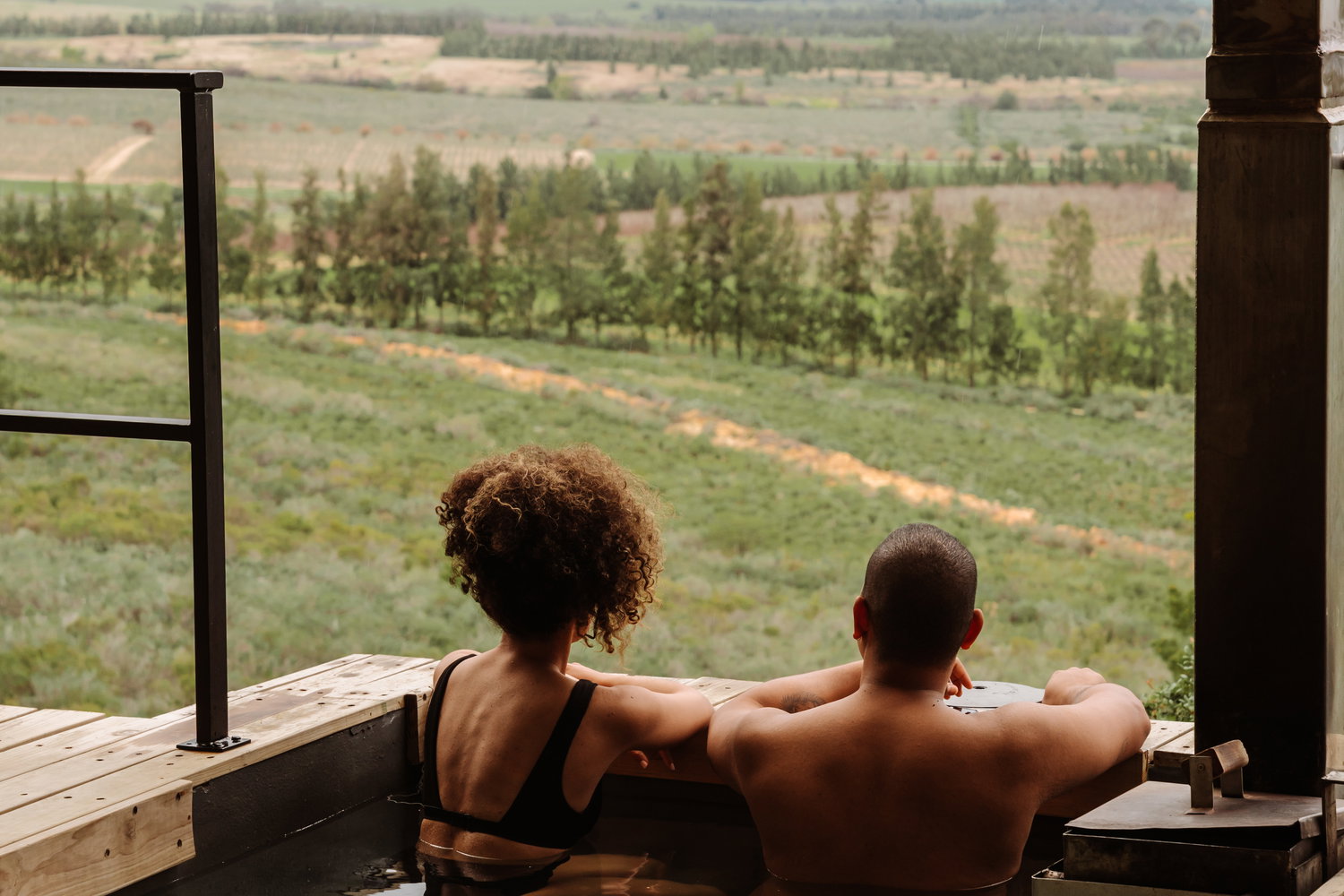The Outpost at Bergsicht Country Cottages: A romantic couple's breakaway in the Tulbagh valley. The image shows a man and woman soaking in a private wood-fired tub—which can be heated for winter or kept cold as a plunge pool in summer—set against a backdrop of rolling vineyards and distant mountains under a dramatic sky.Tulbagh accommodation  Romantic getaway Western Cape  Wood-fired hot tub cottages  Self-catering Tulbagh  Couples retreat South Africa  The Outpost Bergsicht  Wine farm views