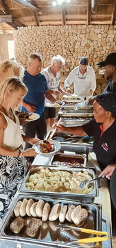 The image captures a group of cruise ship passengers on a shore excursion in Port Elizabeth, eagerly waiting in a short queue to dish up a traditional South African braai (BBQ) lunch. The experience is part of a guided tour, offering visitors an authentic taste of local flavors after an exciting safari or sightseeing adventure. This shore excursion ensures a memorable cultural and culinary experience in South Africa.