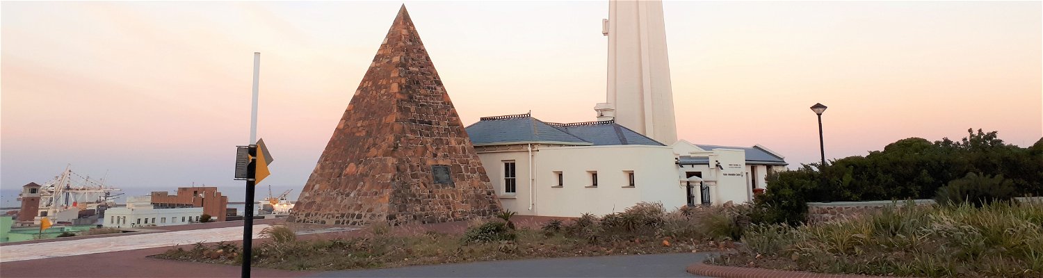 Donkin Reserve in Port Elizabeth, South Africa. This iconic landmark features the striking pyramid-shaped monument, surrounded by lush greenery and overlooking the panoramic views of the city and coastline. The towering lighthouse stands tall against the blue sky, adding to the picturesque scene.