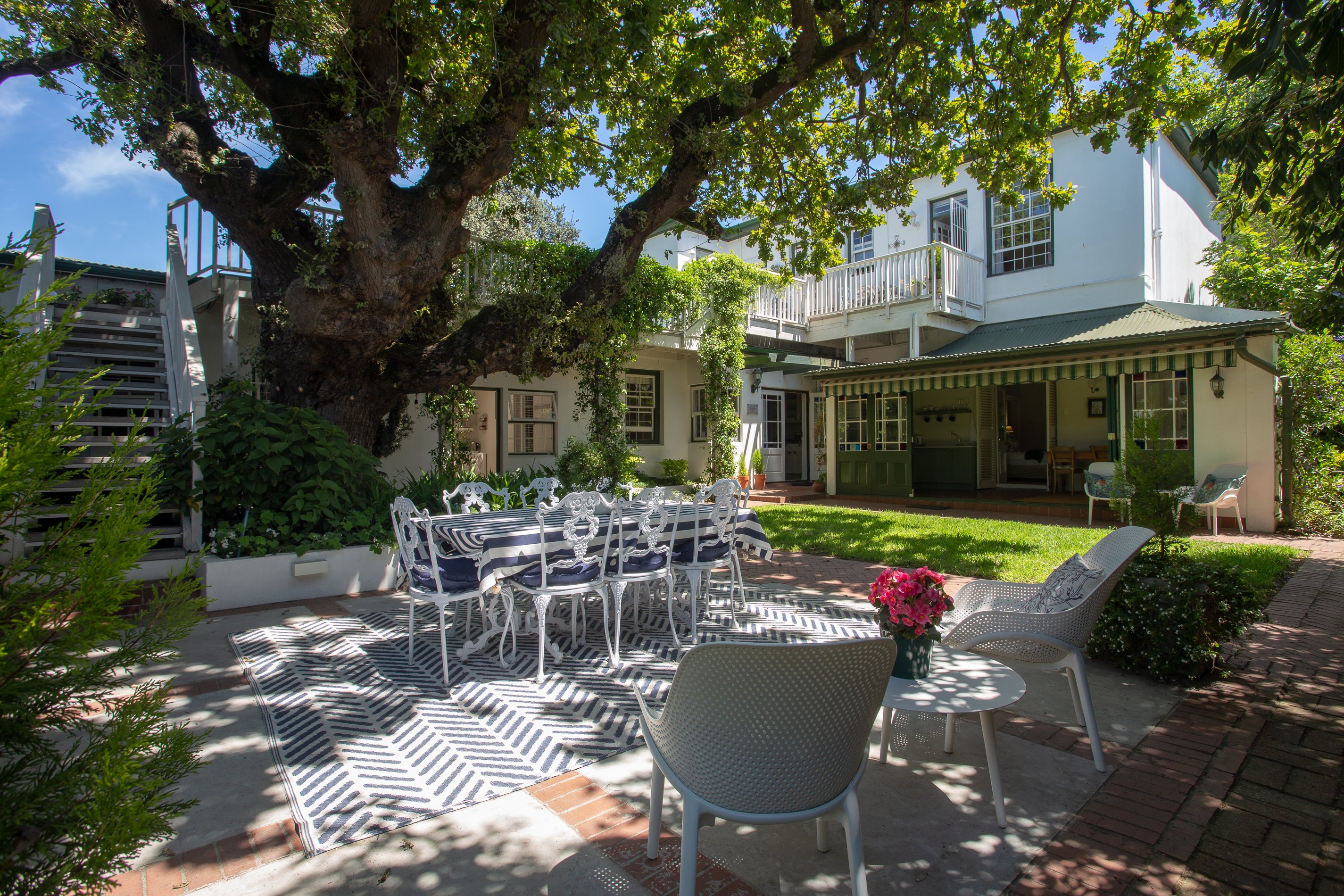 Garden patio with a white metal table and chairs under a large shady tree, beside a white colonial-style guest house.