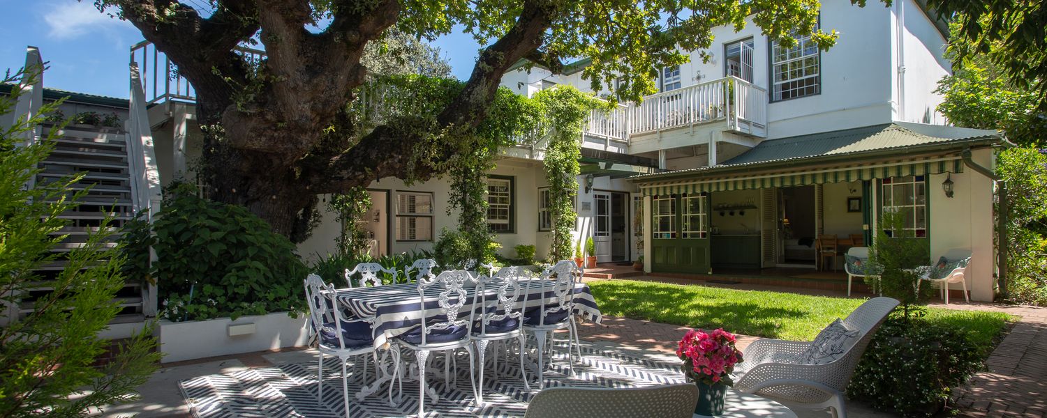 Garden patio with a white metal table and chairs under a large shady tree, beside a white colonial-style guest house.
