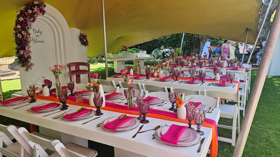 Long tables set with pink napkins, pink-tinted goblets and orange runners under a tent, beside a "Baby in Bloom" floral backdrop.