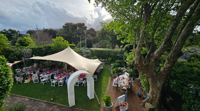 Elevated view of a garden event with long tables set under a beige stretched canopy, with a white arch and a floral sign in the foreground.
