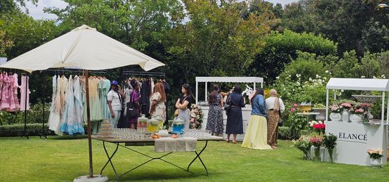 Guests browse pastel dresses on racks and visit a refreshment table beside the white Elancé stall in a sunny garden.