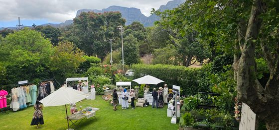 Garden lawn with white umbrellas, clothing racks, and guests browsing market stalls under trees with a mountain behind.