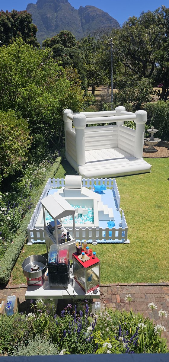 White bouncy castle, ball pit, and candy floss and popcorn stands set up on a lawn in a garden with a mountain behind.