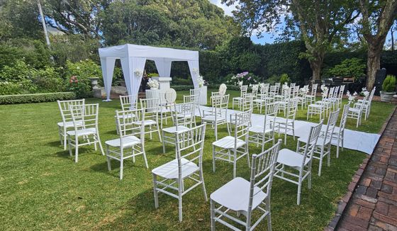 White wedding chairs in rows on a lawn facing a white draped gazebo with floral arrangements, beside a brick path.