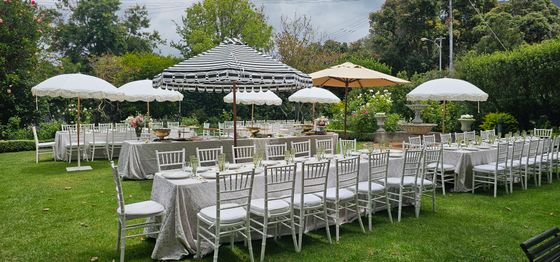 Outdoor garden event space with long tables set with white chairs, grey linens, and striped and cream parasols on green lawn. venue hire rondebosch