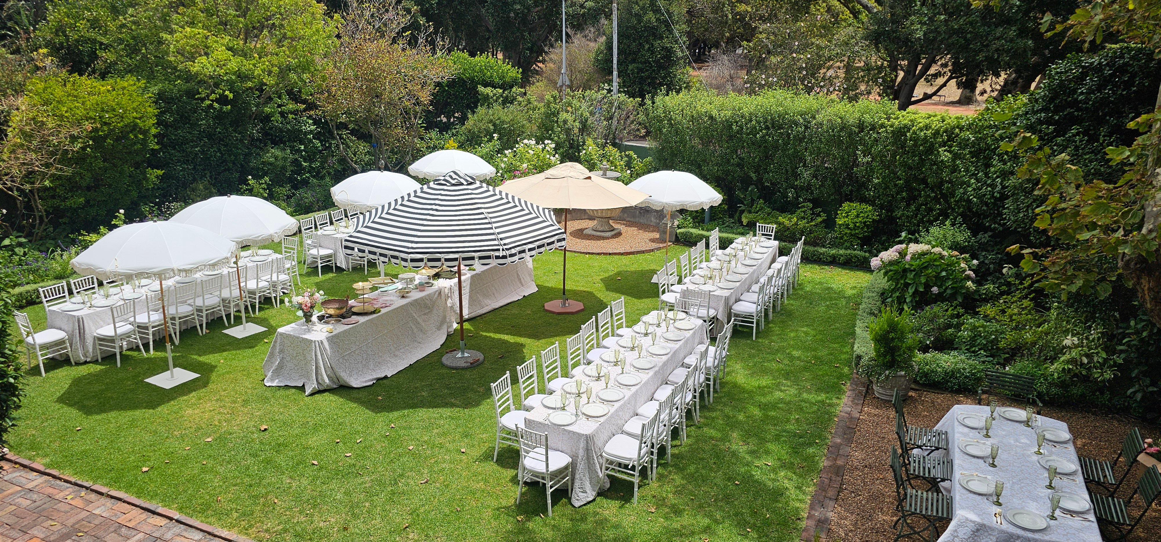 Elevated view of a garden set up with long tables under white and striped umbrellas, dressed for an outdoor event. venue hire rondebosch