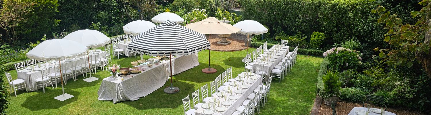Elevated view of a garden set up with long tables under white and striped umbrellas, dressed for an outdoor event. venue hire rondebosch