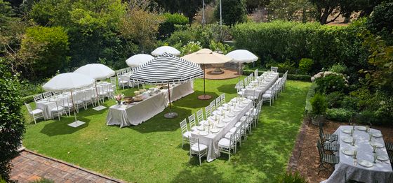 Garden event setup with long tables dressed in white linen, white chiavari chairs, and striped and white parasols on a lawn. venue hire rondebosch