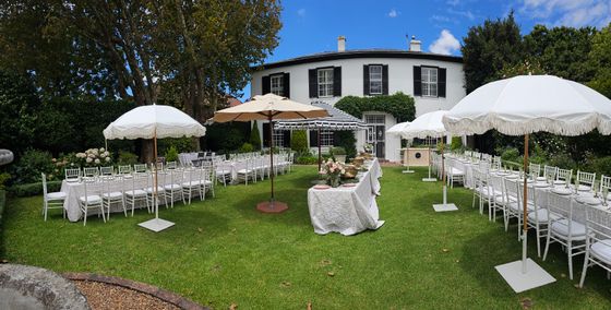 Outdoor garden event set up with white-clothed tables, white chairs, and cream fringed parasols on a lawn in front of a white colonial-style house. venue hire rondebosch