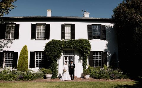 Bride and groom in formal wedding attire stand at the entrance of a white colonial-style house with black shutters and ivy-covered arch. photoshoot at guest house