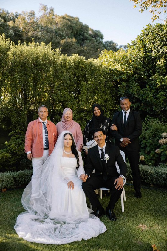 Bride in a white gown and veil, groom in a black suit, and four family members standing in a green garden. venue hire rondebosch