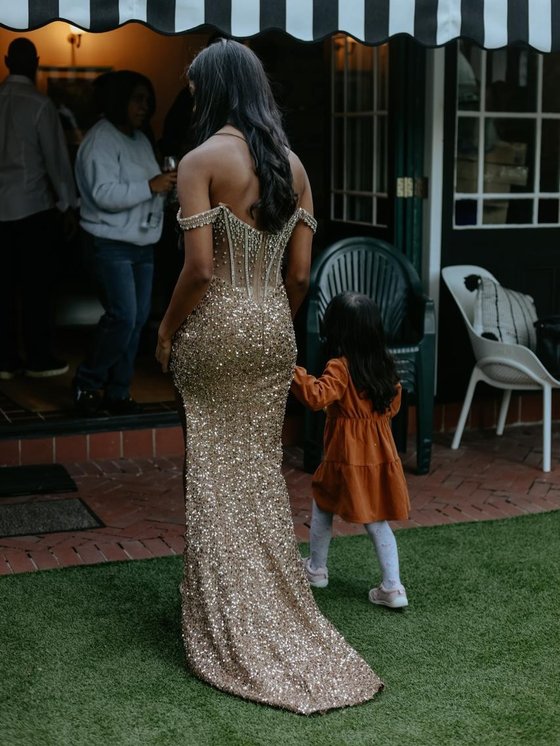 A woman in a sequined gold gown walks across a lawn holding the hand of a young girl in an orange dress.