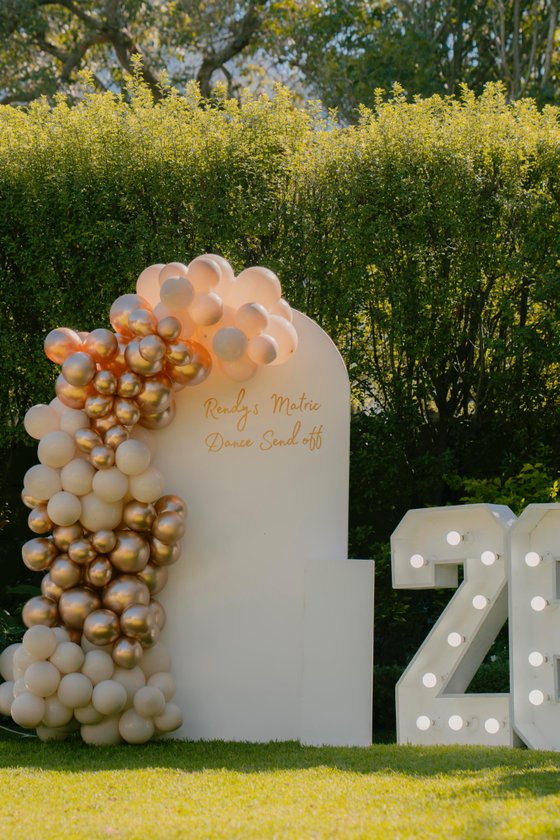 White arch backdrop with gold and cream balloons, gold script reading "Rendy's Matric Dance Send off," and a lit marquee "2" on a lawn.