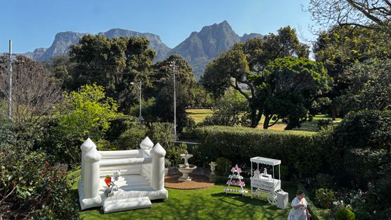 White bouncy castle and a white candy cart on a lawn in a garden, with a mountain range behind. venue hire rondebosch