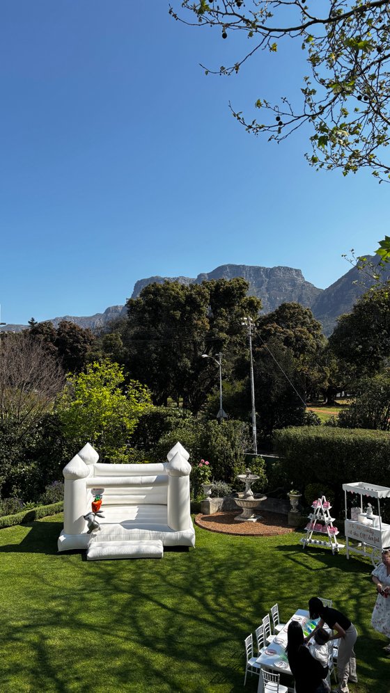 White bouncy castle on a lawn beside a stone fountain, with a dessert table and mountain behind a garden event space.