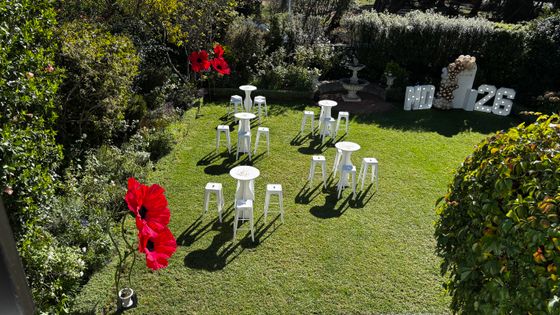 Aerial view of a sunny lawn set with white bistro tables and stools, red poppy-style flowers, and a white "2026" marquee sign.