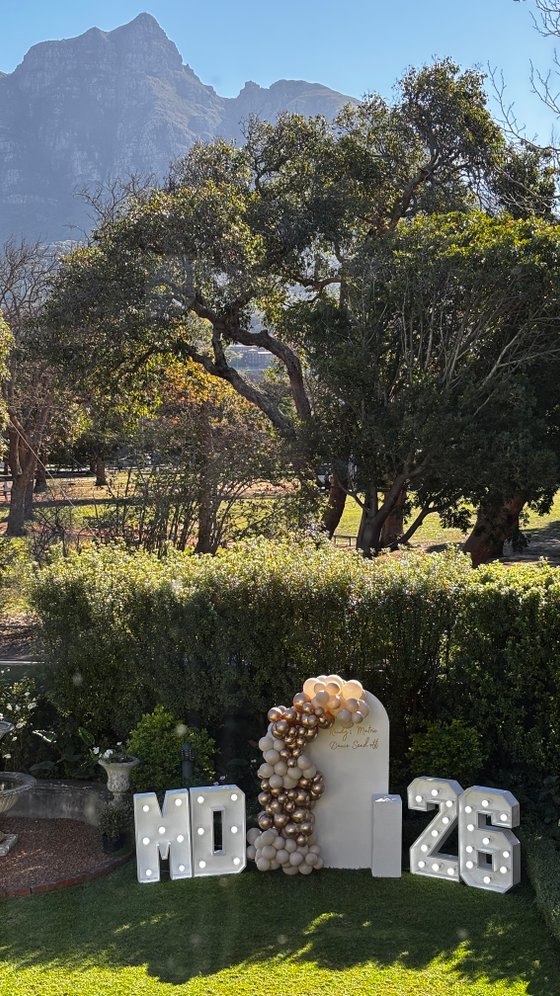 Light-up marquee letters spelling "MR 26" with a gold and cream balloon arch beside a white arched backdrop, set on a lawn with hedges and trees.