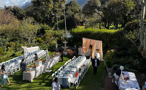 Long tables with white linens, blue place settings and white chairs set up on a lawn in a garden for an outdoor event.