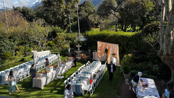 Long garden tables with white linens, blue place settings and white chairs set for a outdoor event on a lawn.