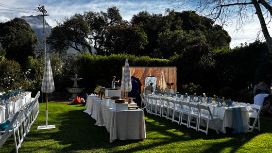 Long garden tables with white linens, blue glassware and white chairs set for an event on a lawn, with a buffet table to the left.