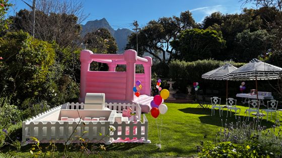 Pink bouncy castle and white picket-fenced play area on a sunny lawn, with striped umbrellas and balloons nearby.