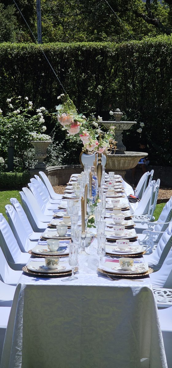 Long garden table set for a formal afternoon tea with white chair covers, teacups, and pink roses under a fountain.