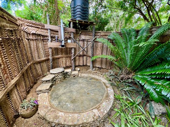 Tatu’s open-air shower at ground level: a round stone floor inside woven palm-leaf walls, under the sky beside a cycad