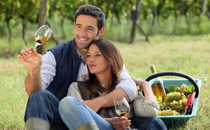 Couple enjoying a romantic wine picnic in a vineyard. The man is holding a glass of white wine up to the light while the woman leans against him, both smiling and relaxed. A picnic basket filled with grapes and a bottle of white wine rests beside them on the grass. Rows of lush green grapevines in the background suggest a serene countryside wine estate, perfect for couples’ getaways, wine tasting tours, or romantic vineyard experiences in South Africa.
