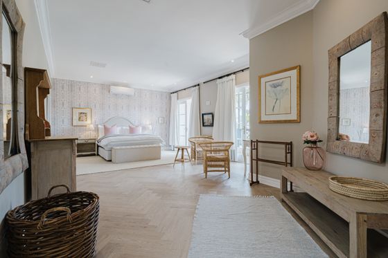 Bedroom with a large white bed with pink pillows, wicker chairs by French doors, and a light herringbone wood floor.