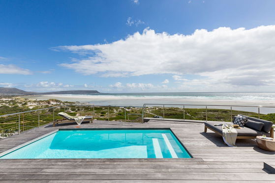 Elevated wooden deck with rectangular turquoise pool at a Cape Beach Villa in Noordhoek, overlooking sandy dunes, a wide shoreline and distant headland, furnished with sun loungers, a daybed and glass railing under a partly cloudy blue sky.