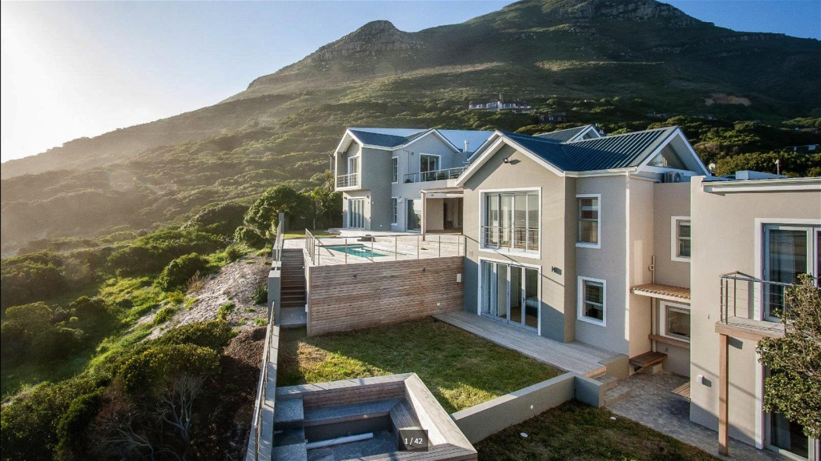 Modern multi-level beige Cape Beach Villa in Noordhoek perched on a hillside with steel-roofed gables, expansive wooden deck with small plunge pool, glass sliding doors and balconies, terraced grassy yard below, rugged green mountainside and sunlit coastal scrubland.