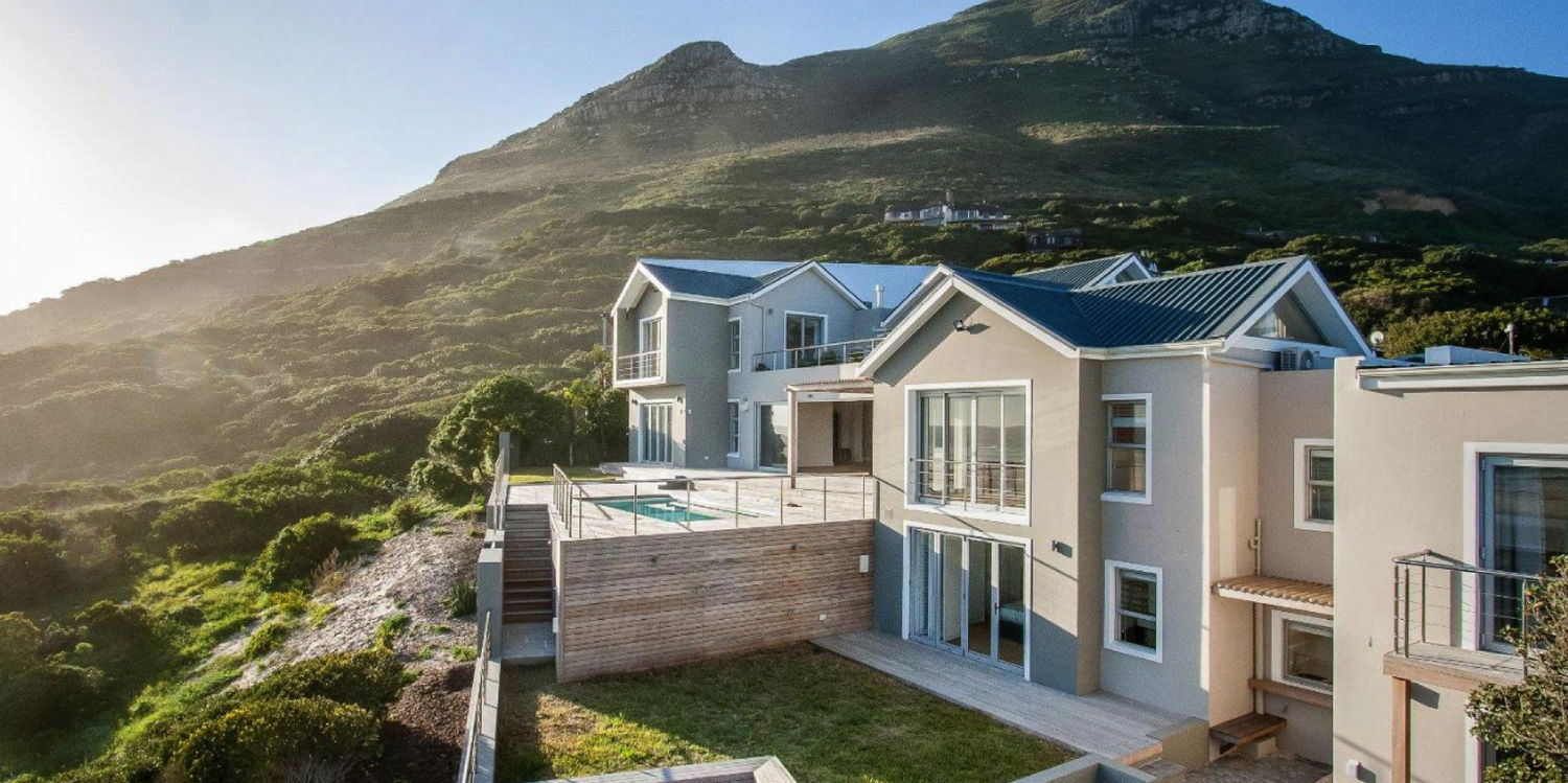 Modern multi-level beige Cape Beach Villa in Noordhoek perched on a hillside with steel-roofed gables, expansive wooden deck with small plunge pool, glass sliding doors and balconies, terraced grassy yard below, rugged green mountainside and sunlit coastal scrubland.
