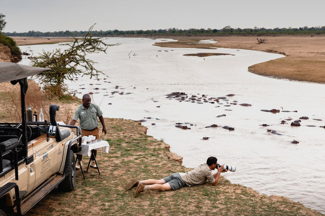 male guest taking photos lying down on water edge looking at hippos by Chichele Presidential Camp Zambia