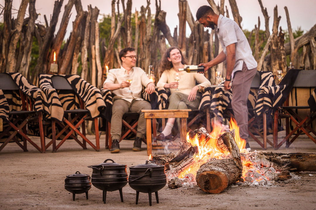 couple sitting in fireplace boma by fire with food pots in front at Jao Camp Wilderness Okavango Delta Botswana