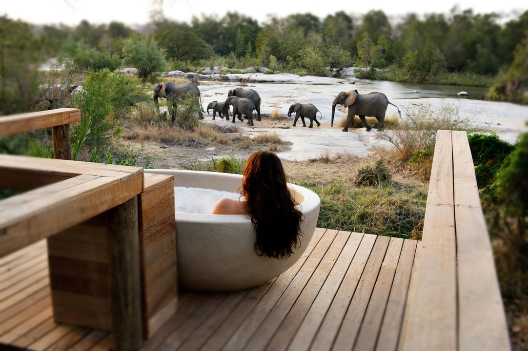 lady in bathtub on deck of granite suites looking at elephant in river by Lodolozi