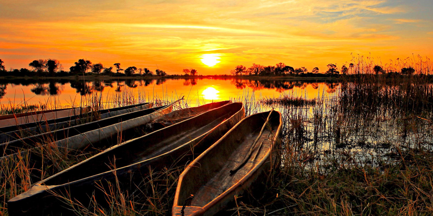 sun setting over Okavango delta with mokoros on riverbank in foreground