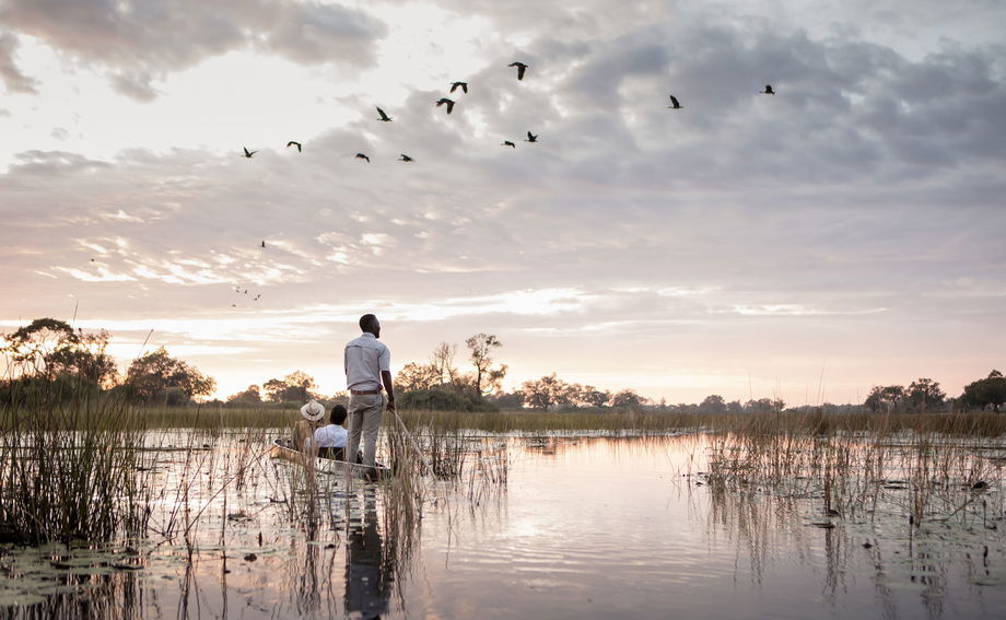 guest and guide in mokoro with birds flying overhead at Vumbura Plains Camp Wilderness Okavango Delta