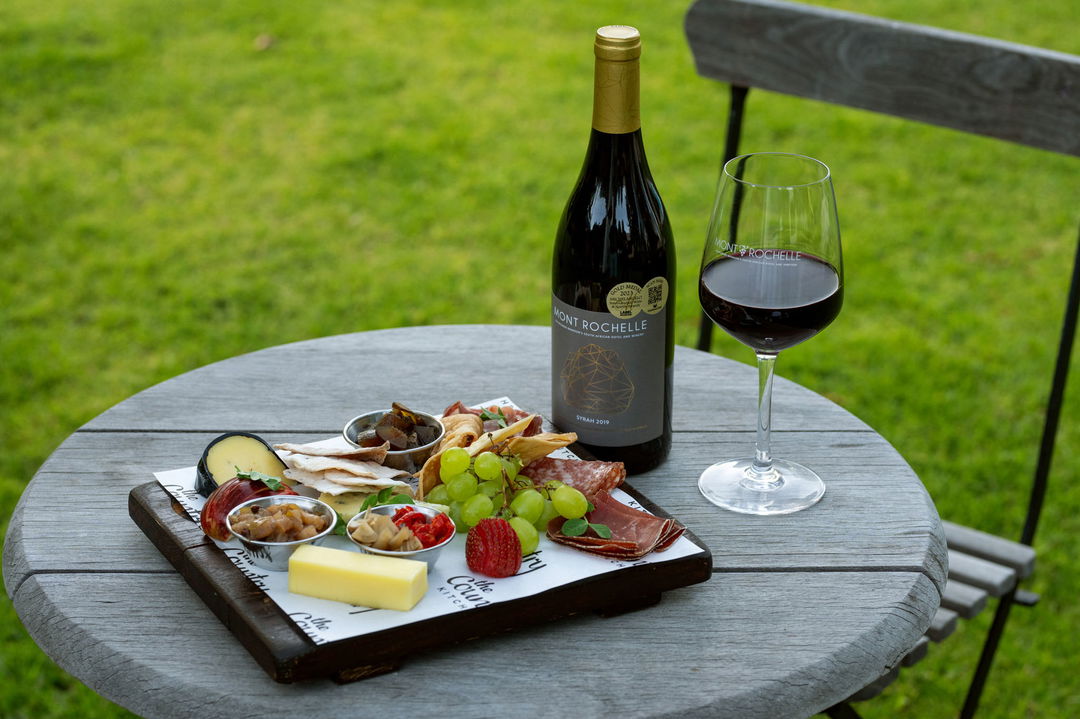 cheese and wine platter and red wine bottle on a table at exterior dusk view of the illuminated facilities of Mont Rochelle Wine Estate