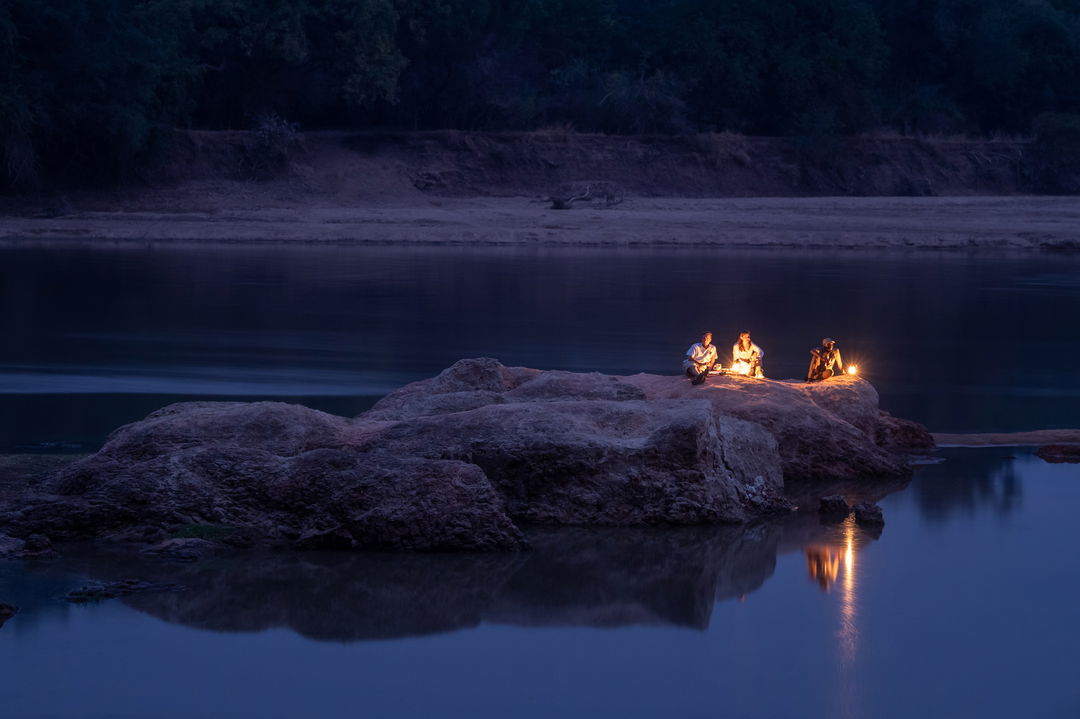 guests sitting by lantern light at night on outcrop of river by Puku Ridge Camp Zambia
