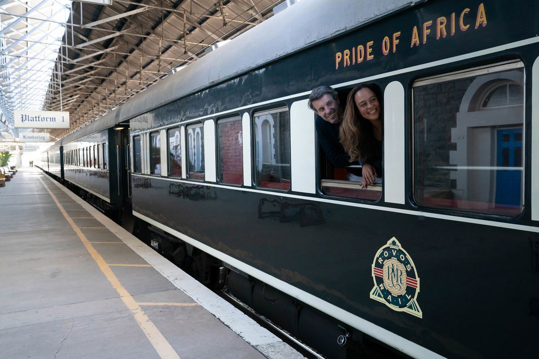 side view of some carts of the train with passengers looking out the window on station platform at Rovos rail luxury train safaris