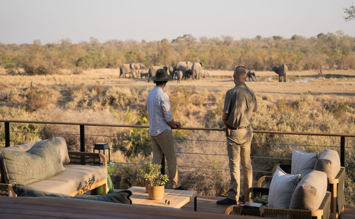 Two guests standing on deck of Thornybush Simbambili lodge looking at herd of elephants by waterhole in background